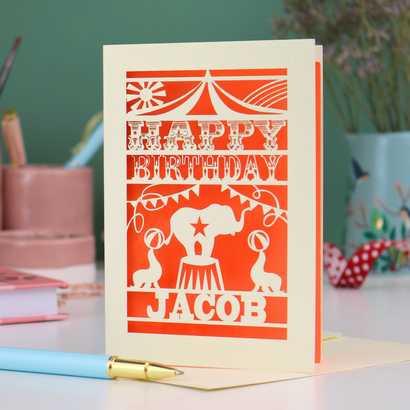 A laser cut personalised birthday card with circus design and text "Happy Birthday Jacob", in cream with an orange paper insert, on white desk with stationery items