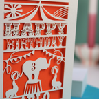 A laser cut birthday card with circus big top design, "Happy Birthday" and a name, with an age cut out.