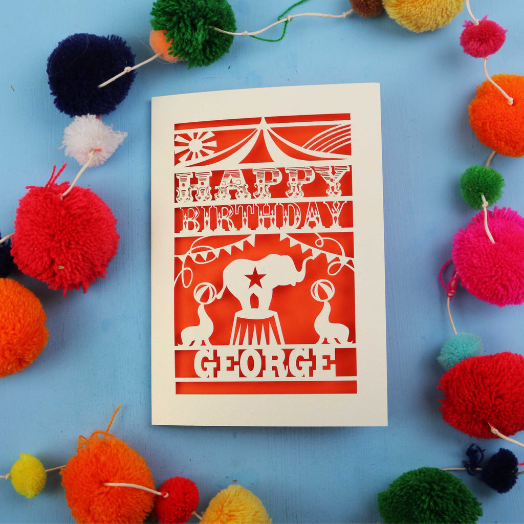 Personalised cut out Circus Birthday card with 'Happy Birthday George' text on a blue background with colourful pom-poms.