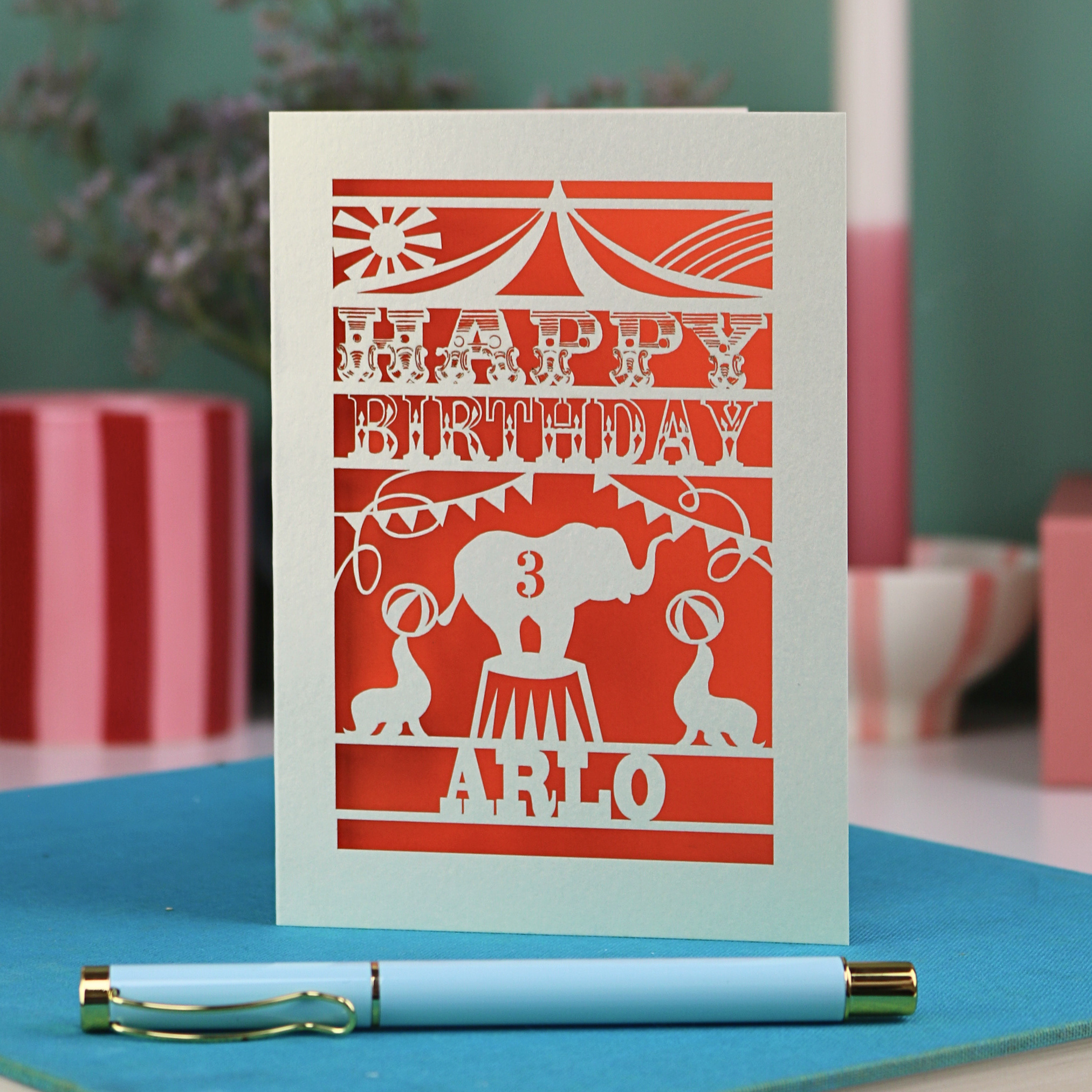 A laser cut birthday card with circus big top design, "Happy Birthday" and a name, with an age cut out.