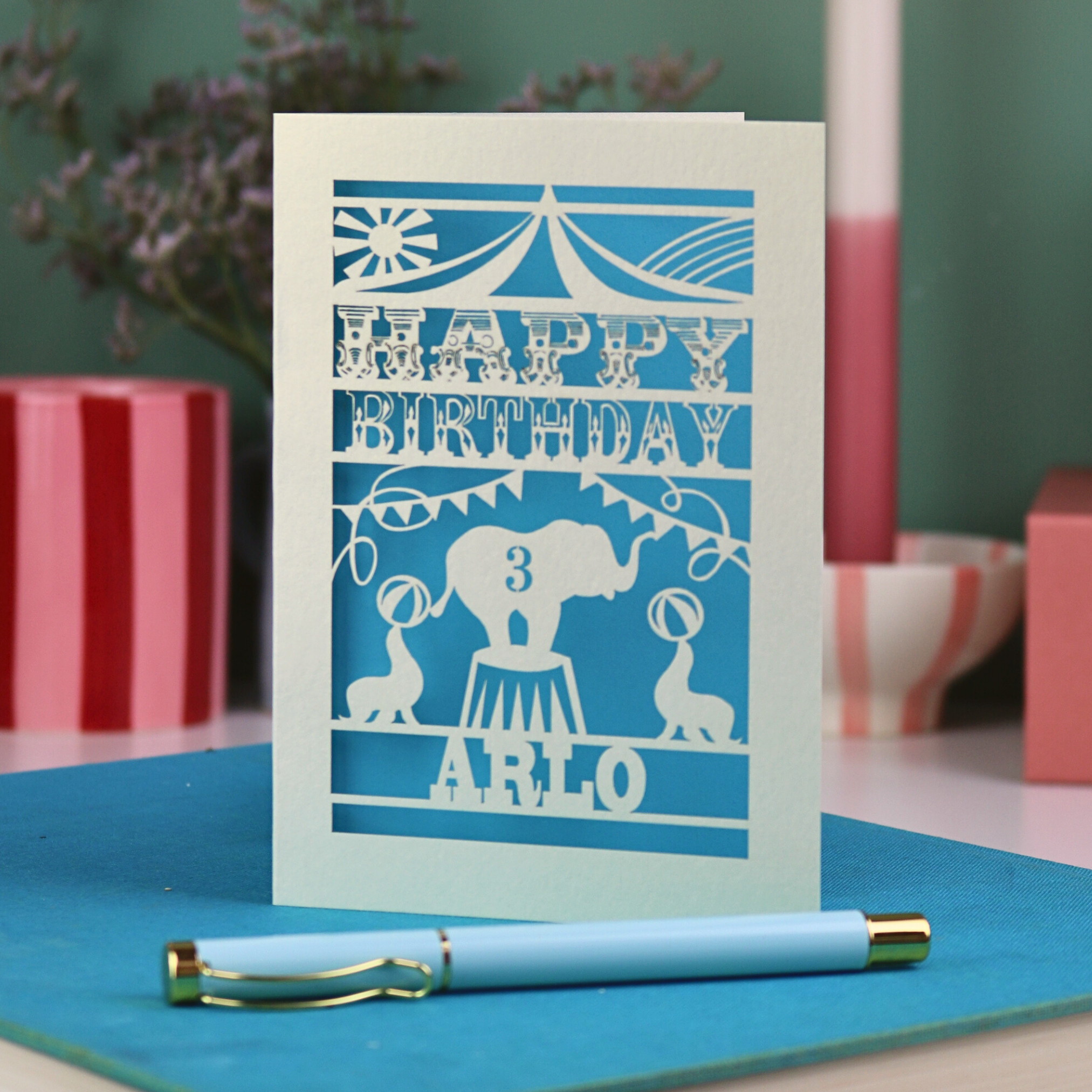 A laser cut birthday card with circus big top design, "Happy Birthday" and a name, with an age cut out.