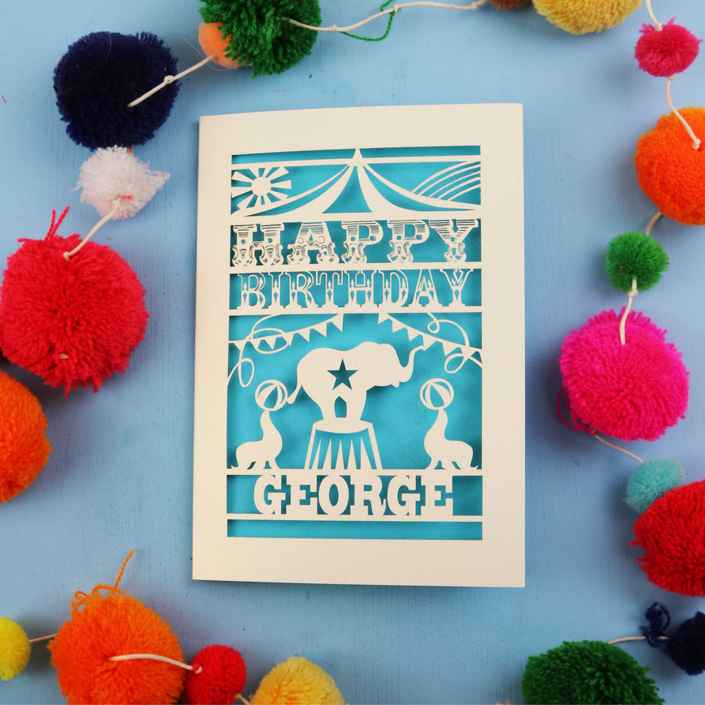 Personalised kids' Circus Birthday card with 'Happy Birthday George' text on a blue background with colourful pom-poms.