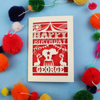 Personalised paper cut Circus Birthday card with 'Happy Birthday George' text on a blue background with colourful pom-poms.