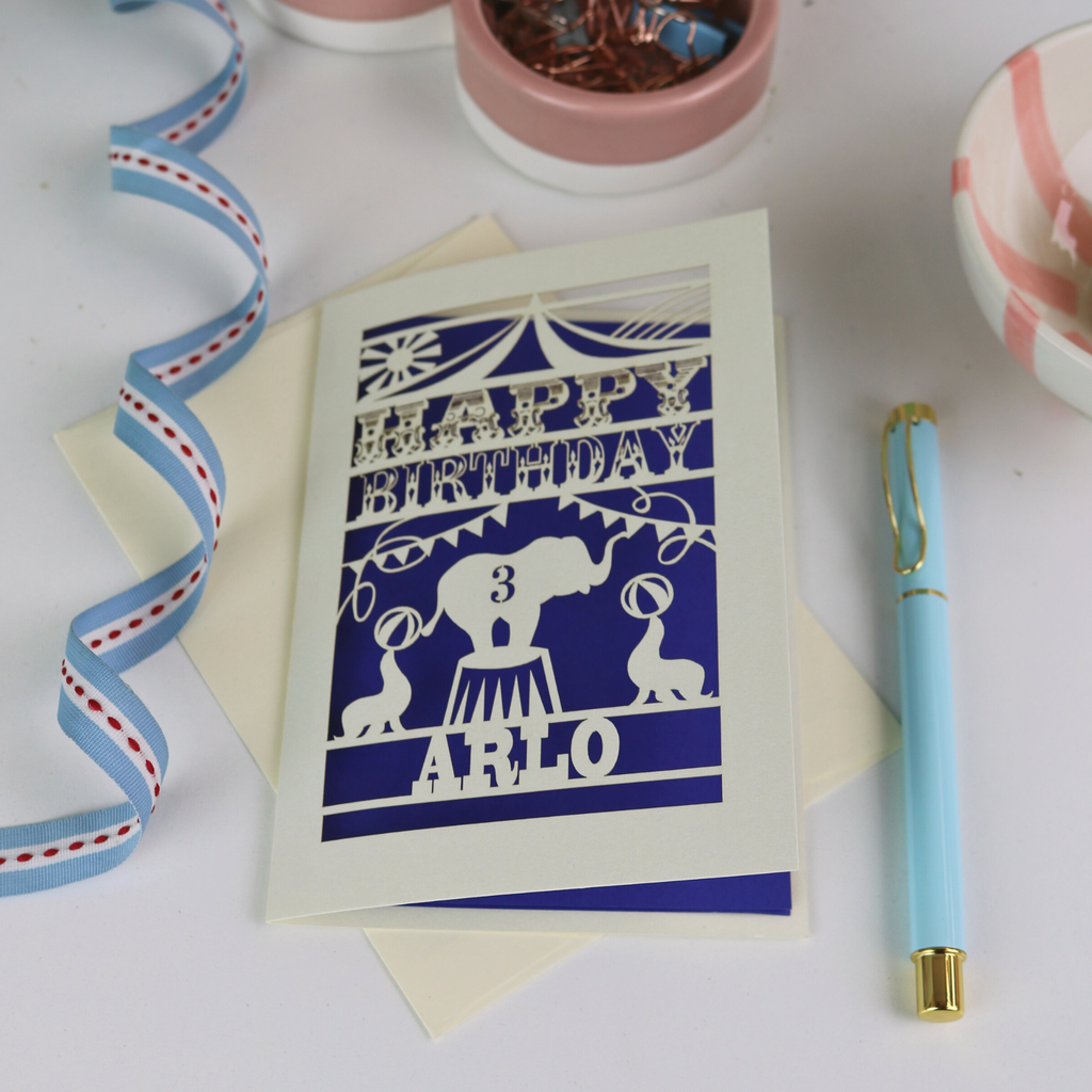 A laser cut birthday card with circus big top design, "Happy Birthday" and a name, with an age cut out.