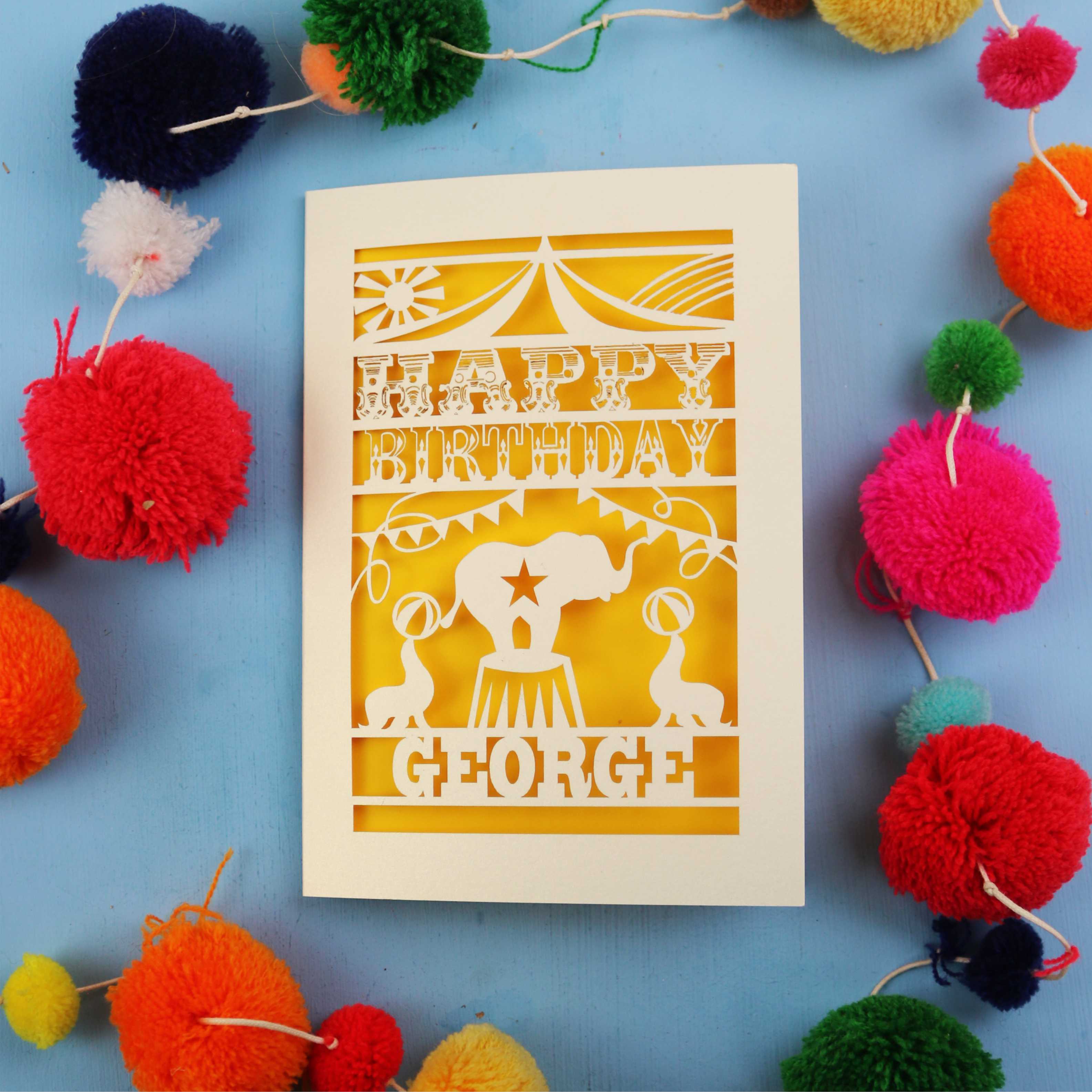 Personalised Colourful Circus Birthday card with 'Happy Birthday George' text on a blue background with colourful pom-poms.