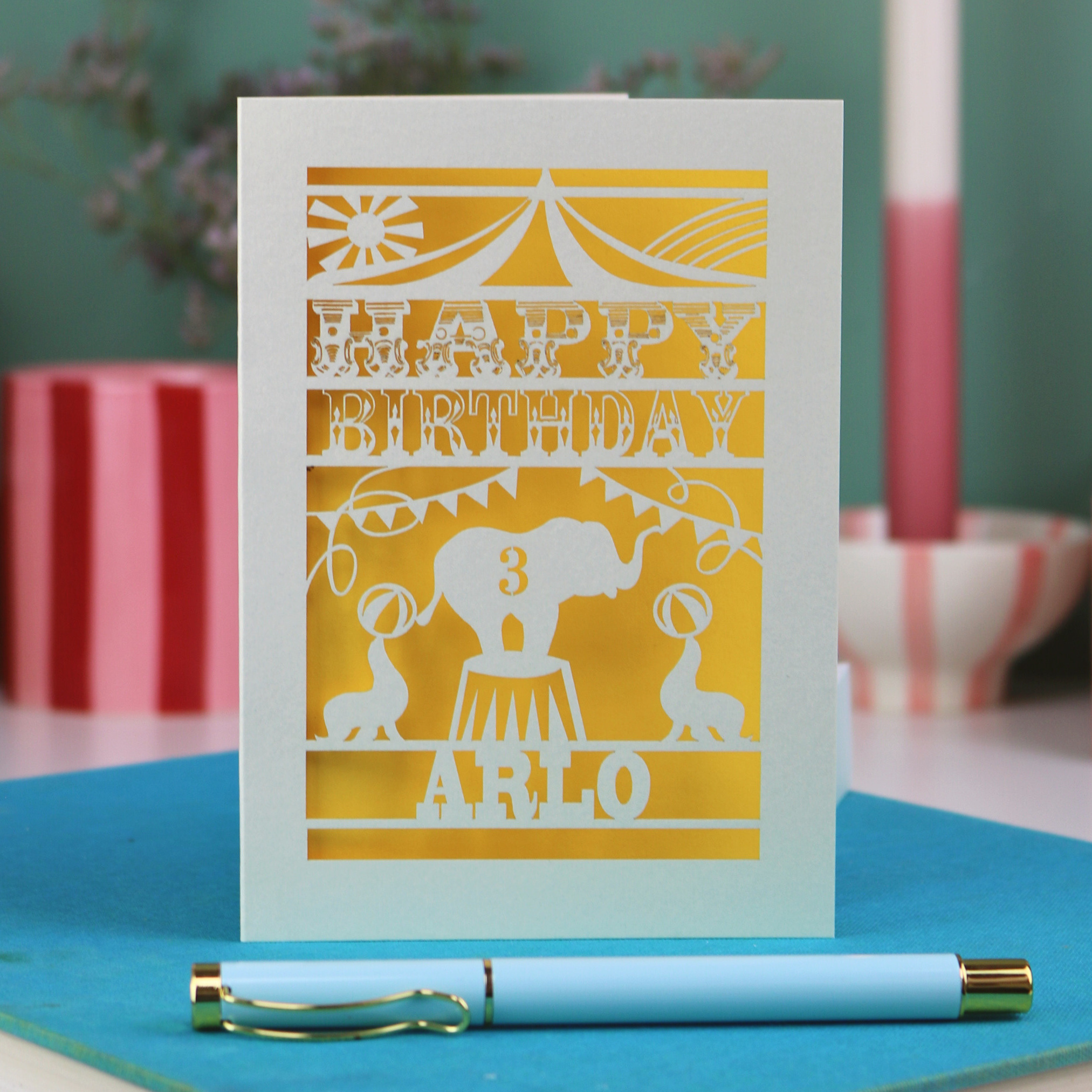 A laser cut birthday card with circus big top design, "Happy Birthday" and a name, with an age cut out.