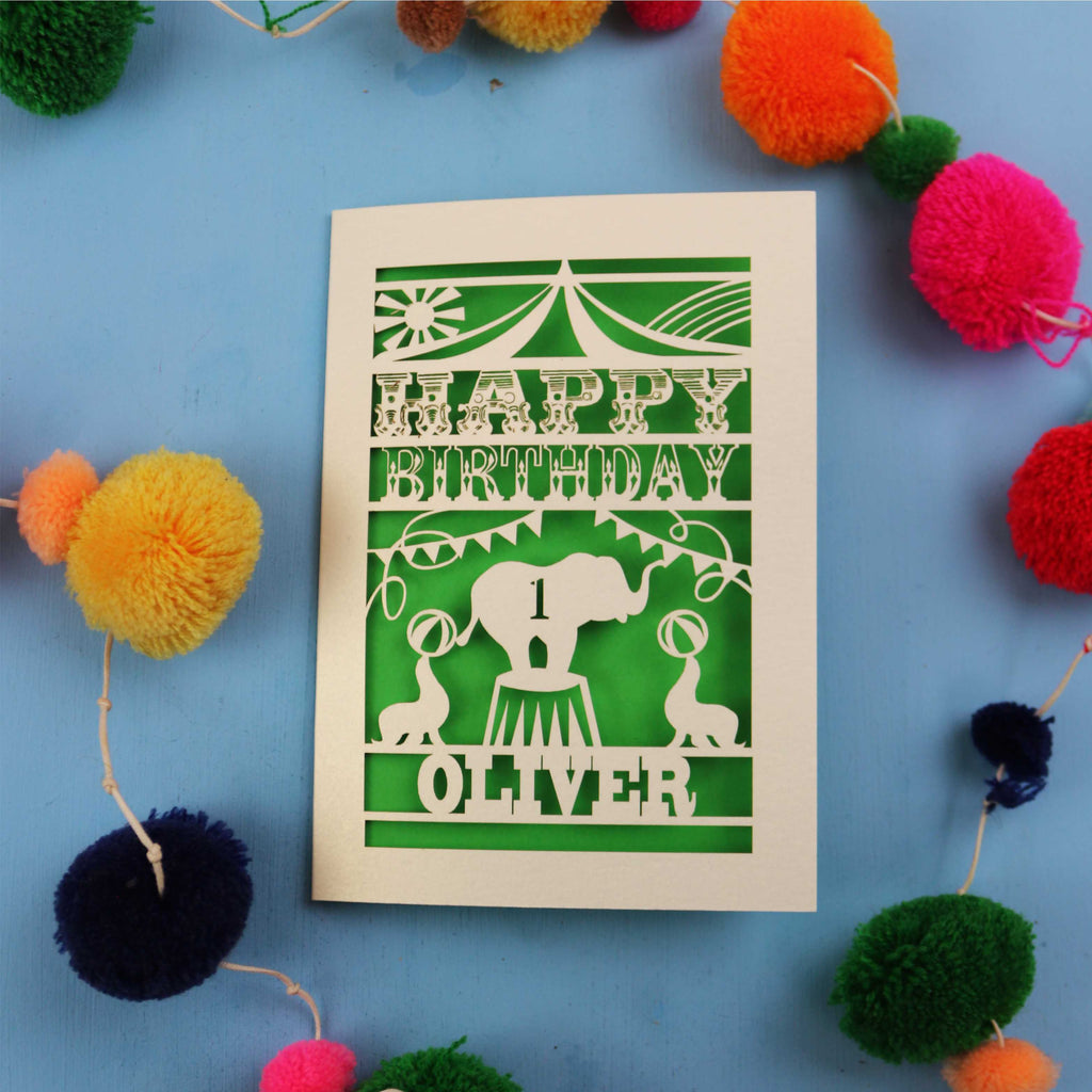 Paper cut First Birthday card with cut-out circus design on a blue background with colourful pom-poms