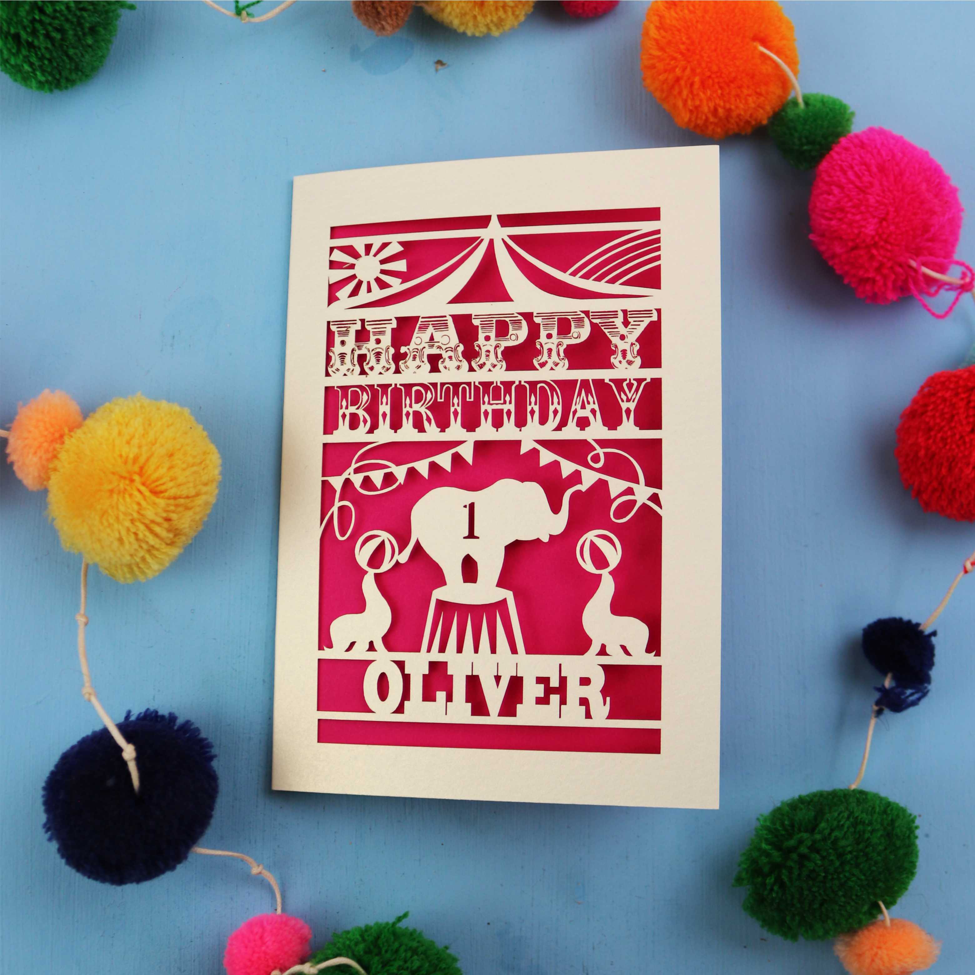 Paper cut First Birthday card with cut-out circus design on a blue background with colourful pom-poms
