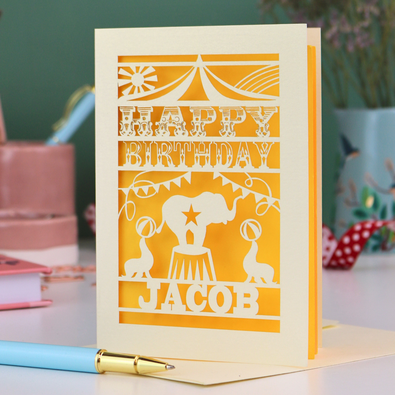 A laser cut personalised birthday card with circus design and text "Happy Birthday Jacob", in cream with a yellow paper insert, on white desk with stationery items