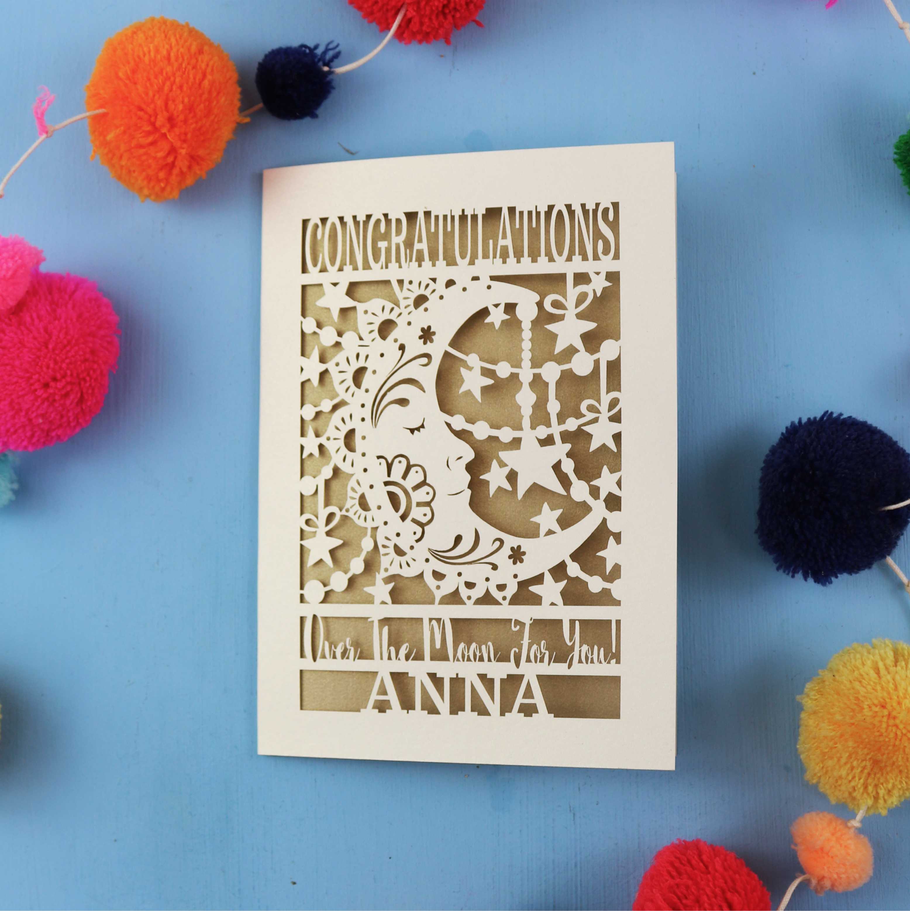Decorative congratulations card with cut-out design on a blue background with colourful pom-poms.