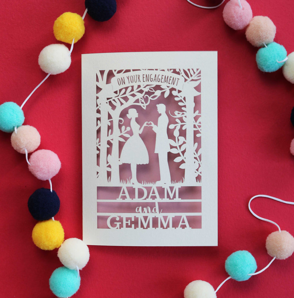A personalised paper cut engagement card with silhouettes of a couple making a heart shape with their hands. Paper cut card says "On your engagement" and has their names under the couple