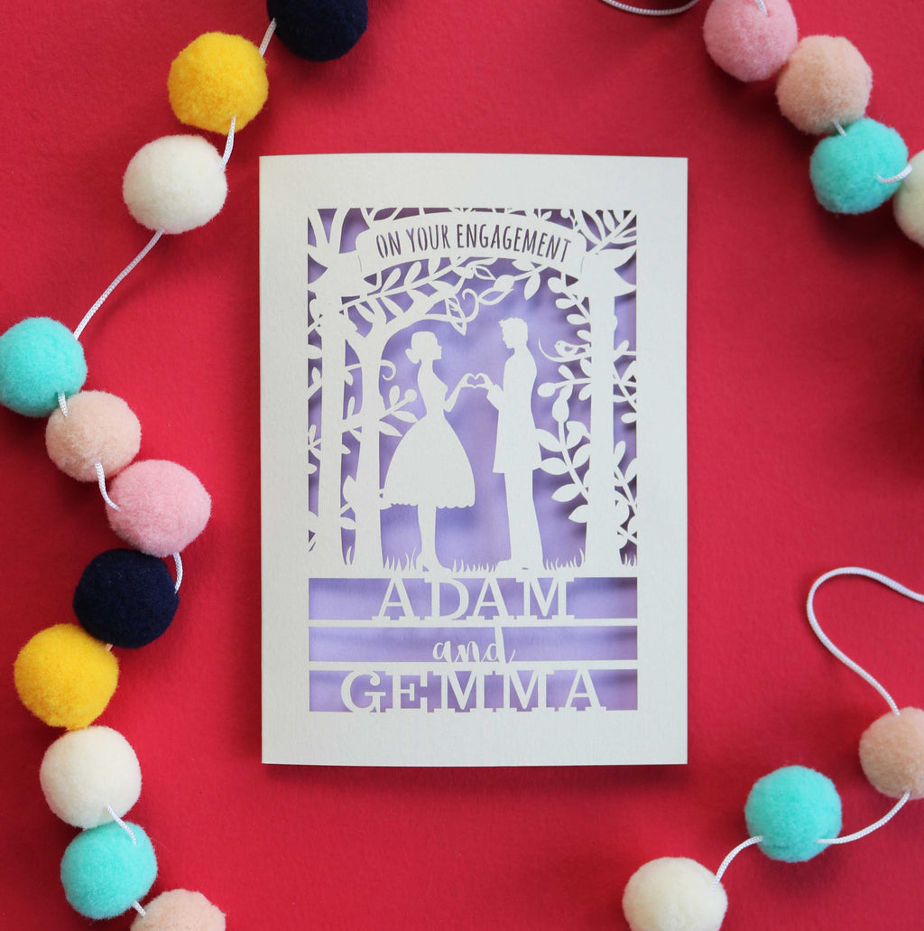 A personalised engagement card, laser cut from cream card with a lilac paper backing. Engagement card features two people, a man and a woman ctanding and making a heart with their hands. 