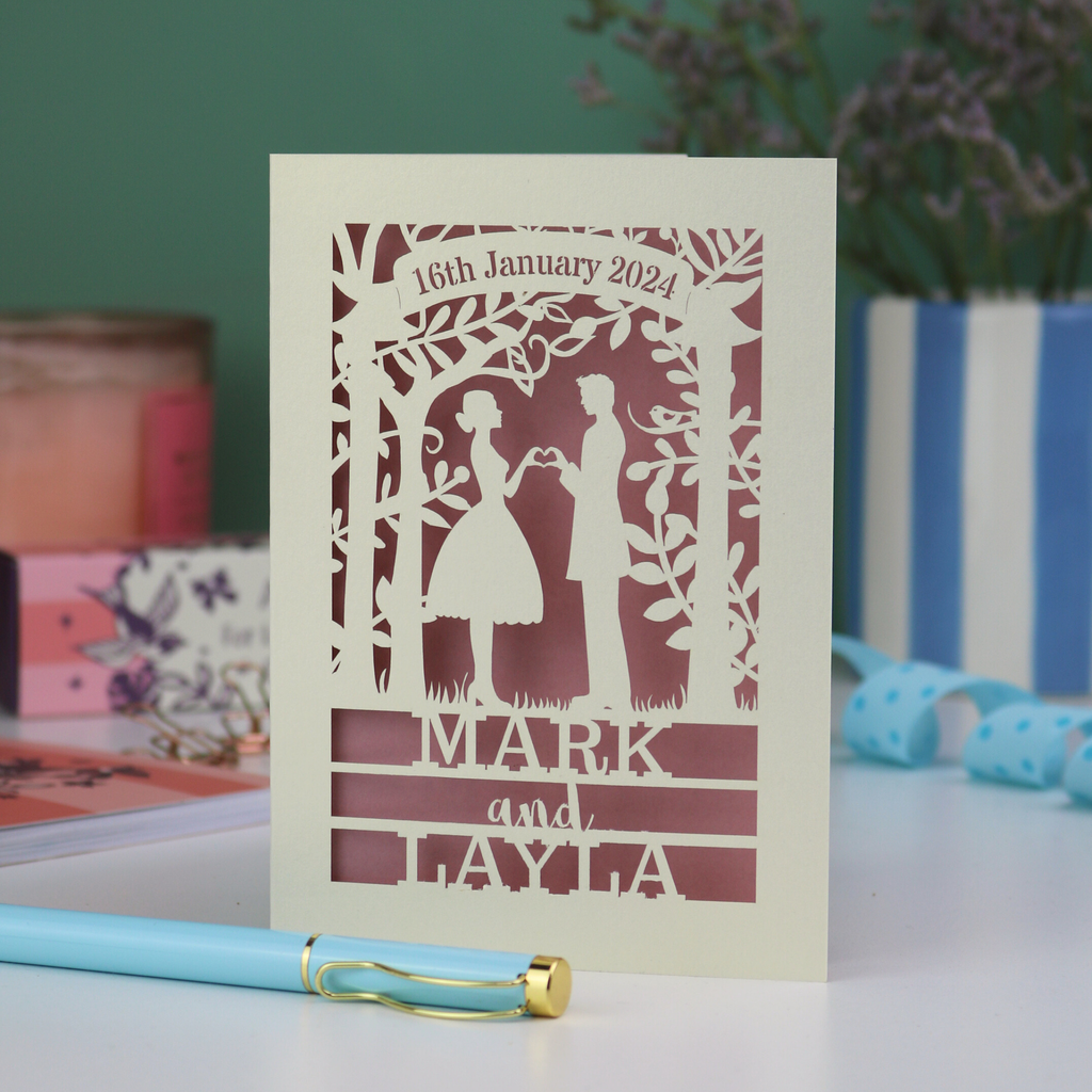 Personalized wedding card with laser-cut design of a couple, date, and names on a table with stationery items.