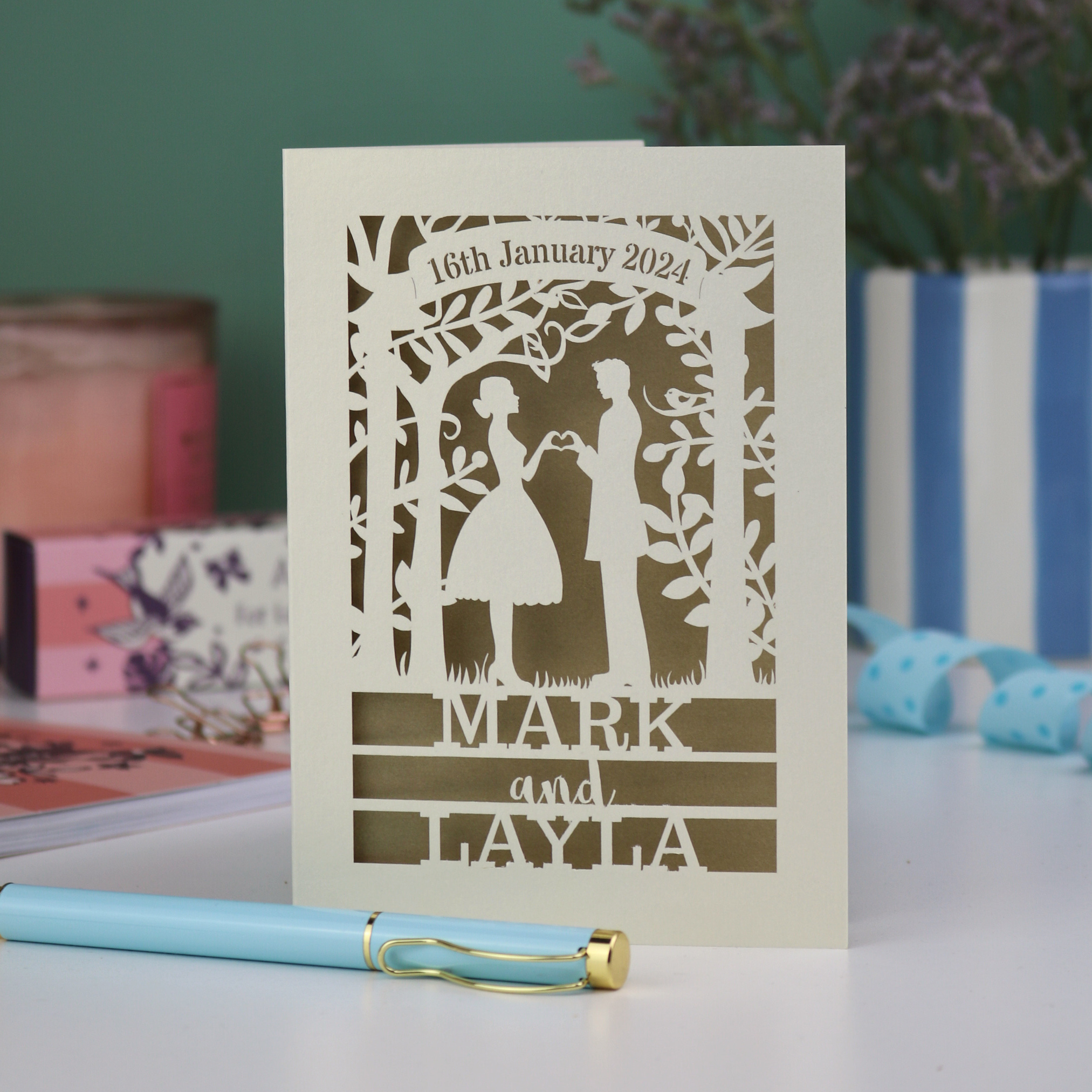 Personalized wedding card with laser-cut design of a couple, date, and names, placed on a surface with a pen and decorative items in the background.