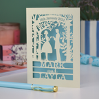 Personalized wedding card with laser-cut design of a couple, date, and names on a light blue background.
