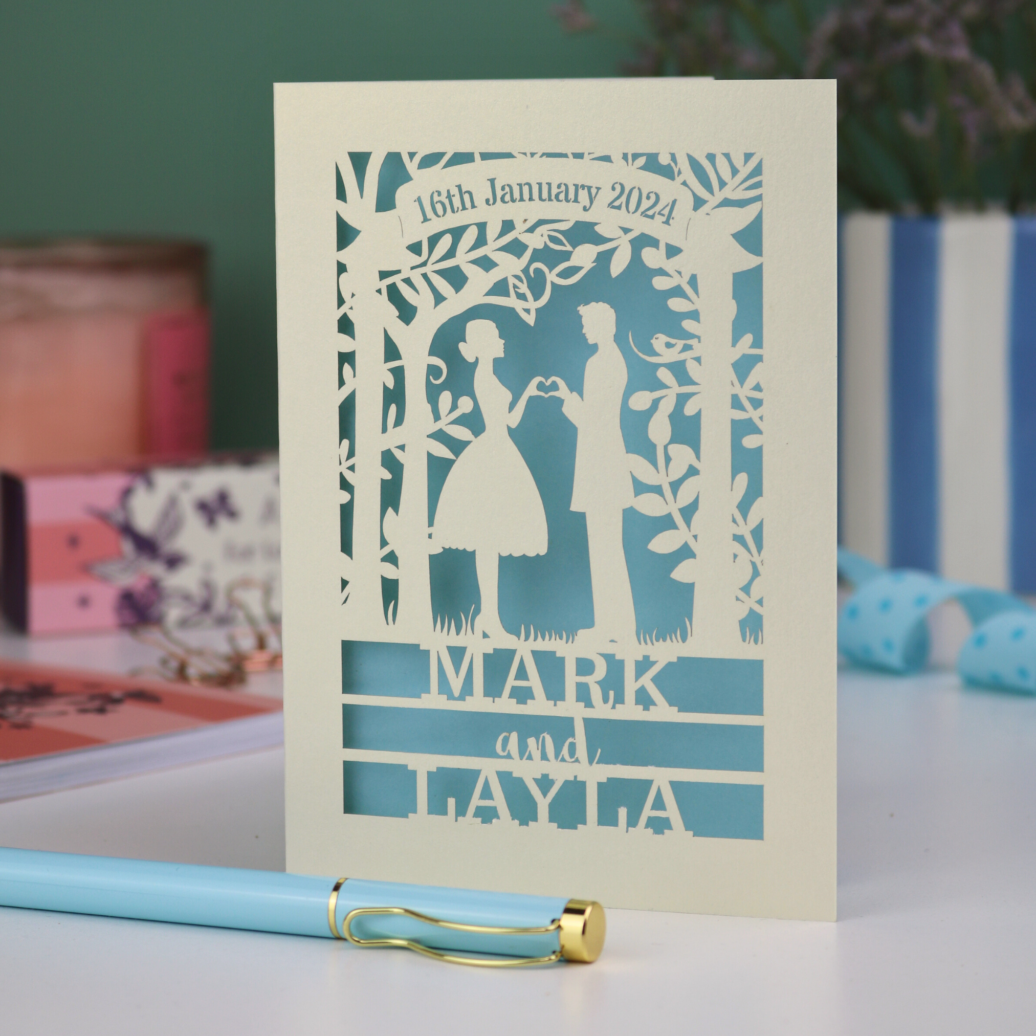 Personalized wedding card with laser-cut design of a couple, date, and names on a light blue background.