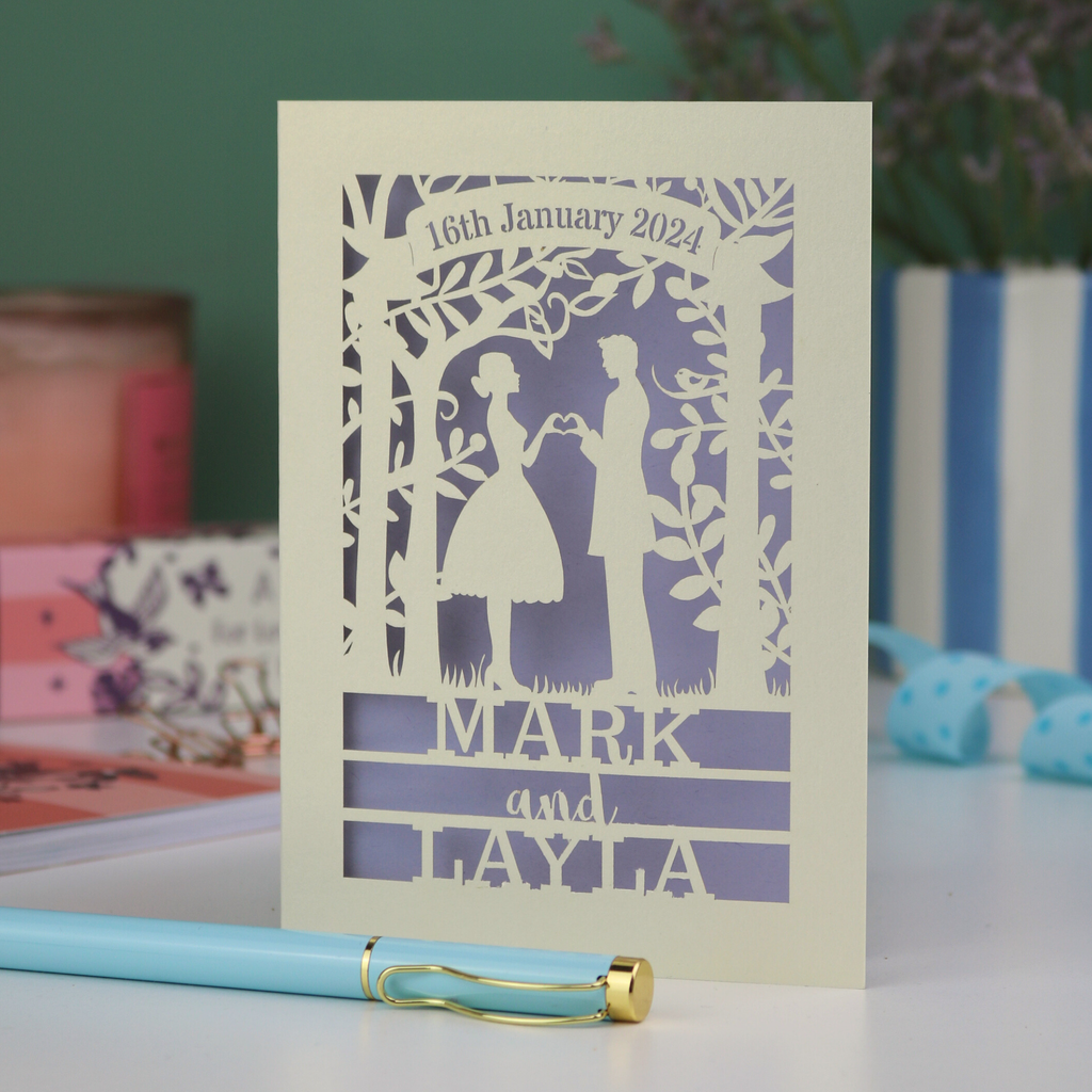 Personalized wedding card with laser-cut design of a couple, date, and names on a table with stationery items.
