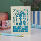 Personalized wedding card with cut-out design of a couple, date, and names on a table with stationery items.