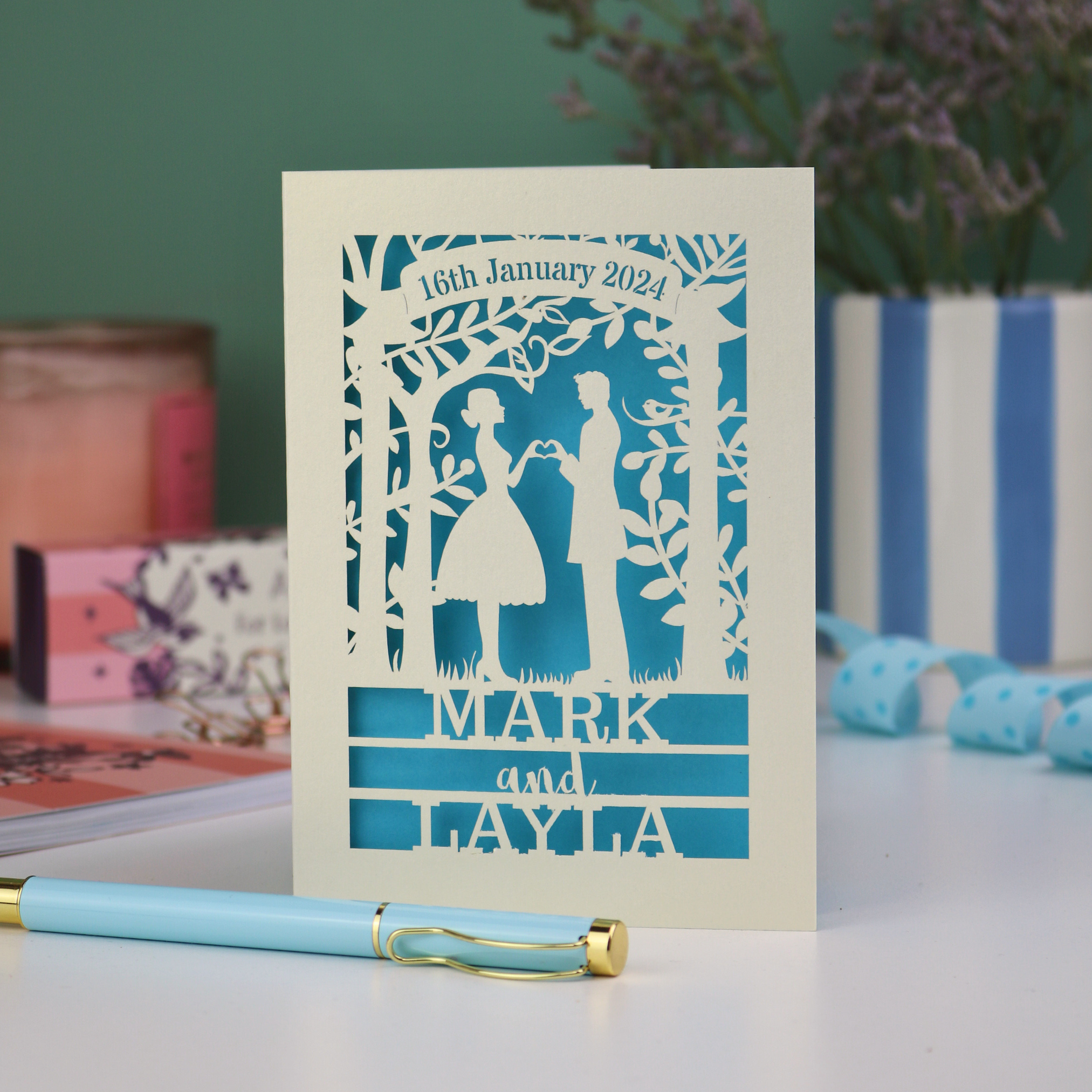 Personalized wedding card with cut-out design of a couple, date, and names on a table with stationery items.