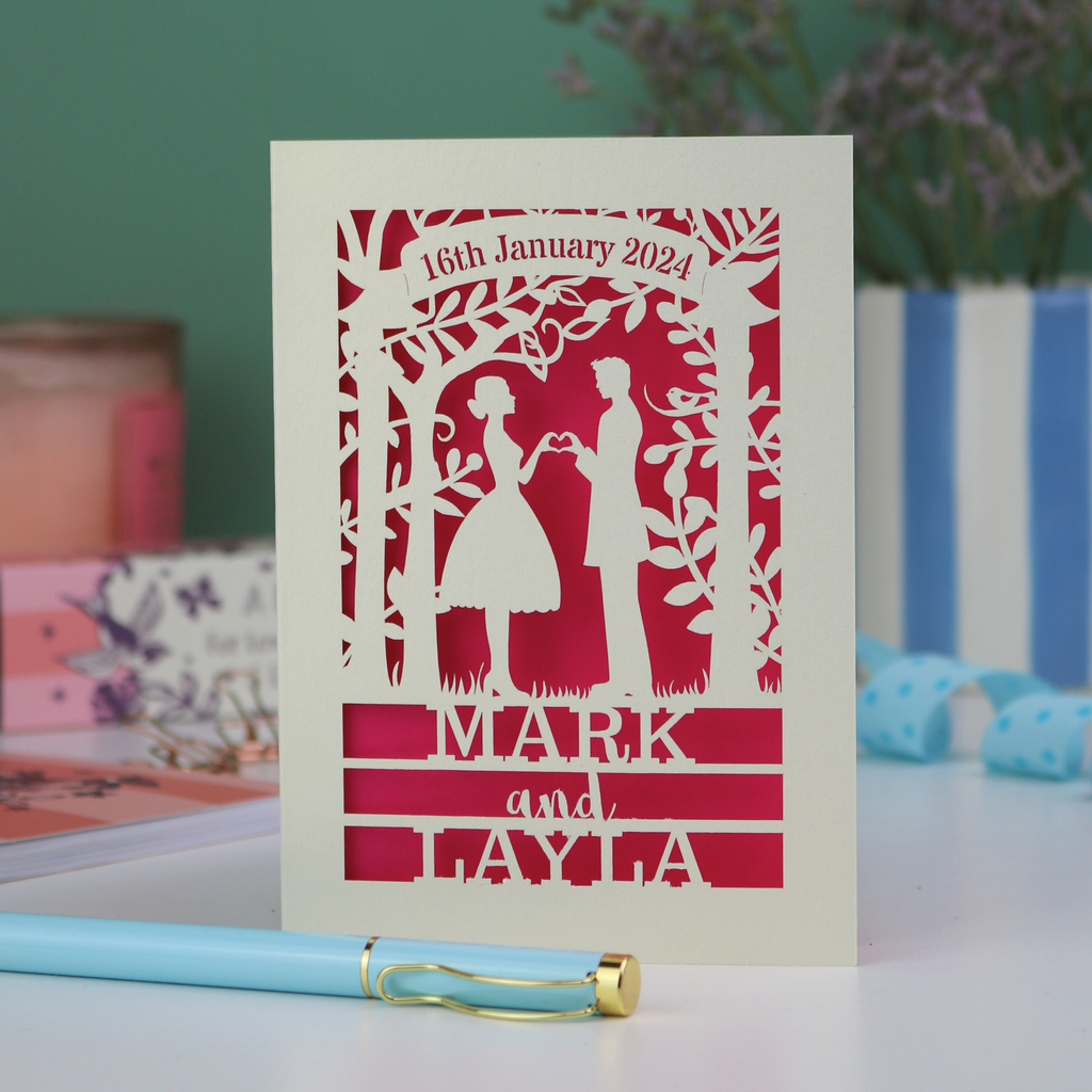 Personalised wedding card with cut-out design of a couple, date, and names on a table with a pen and decorative items.