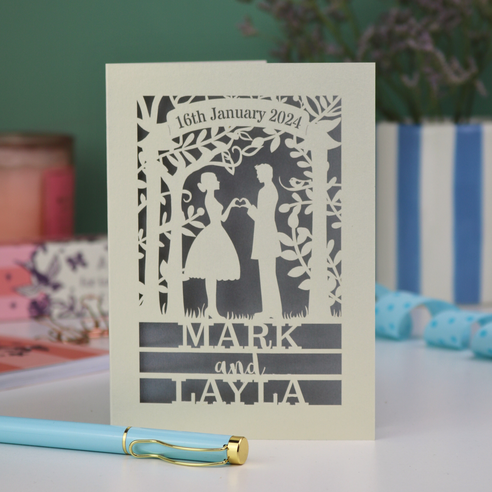 Personalized wedding card with cut-out design of a couple, date, and names on a table with decorative items.
