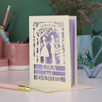 Personalised wedding card with laser-cut design of a couple, names, and date on a desk with stationery items.