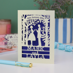 Wedding invitation card with cut-out design of a couple, date, and names on a white surface with decorative items.