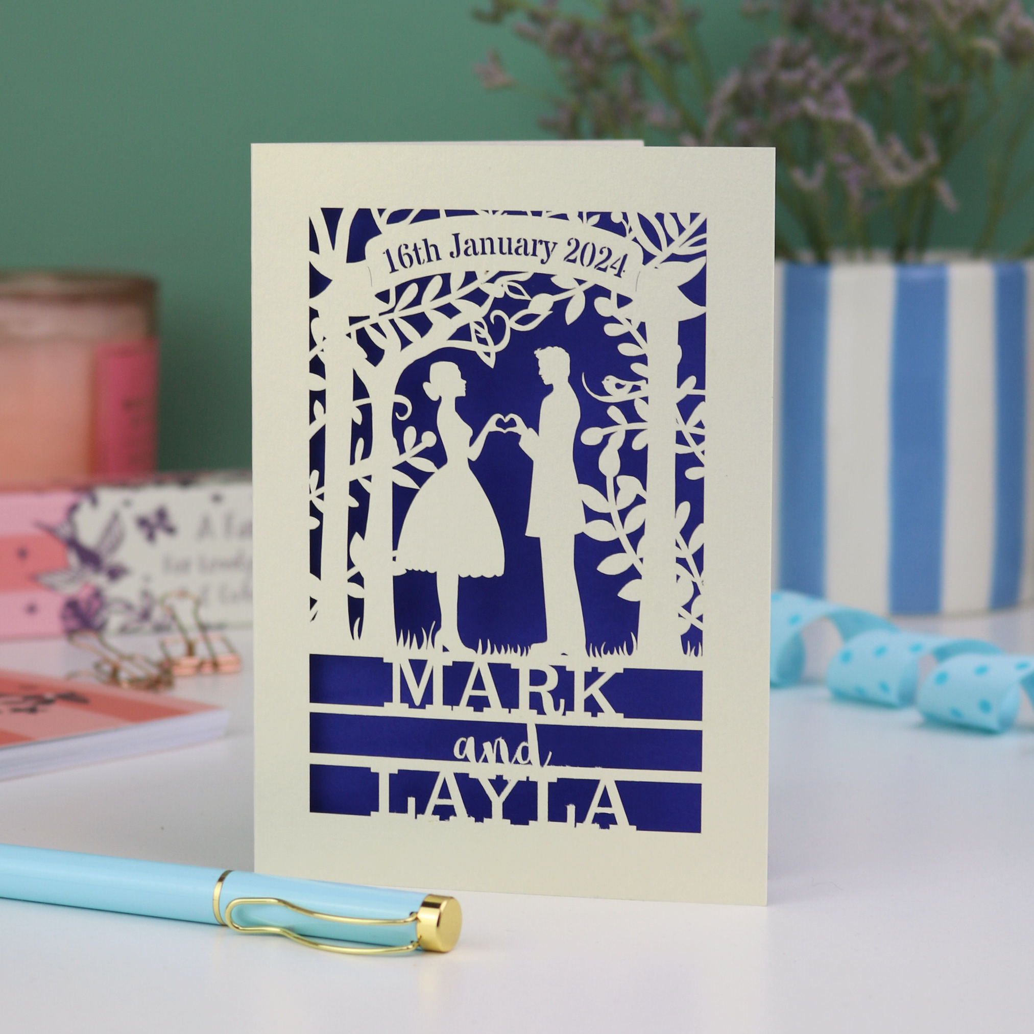 Wedding invitation card with cut-out design of a couple, date, and names on a white surface with decorative items.