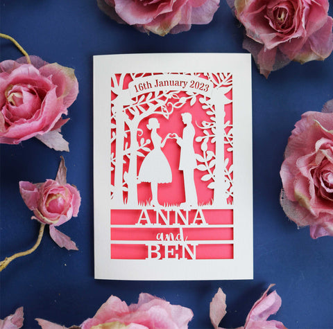 Laser cut wedding cards personalised and made to order. A laser cut wedding card that features the silhouettes of a couple, and is personalised with a date in a banner at the top and the names of the couple over three lines of text at the bottom. - A6 (small) / Coral