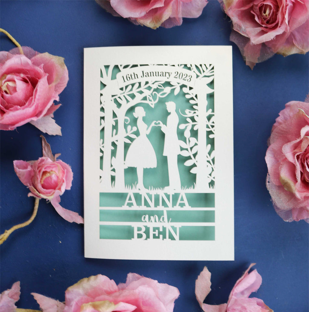 A laser cut wedding card that features the silhouettes of a couple, and is personalised with a date in a banner at the top and the names of the couple over three lines of text at the bottom