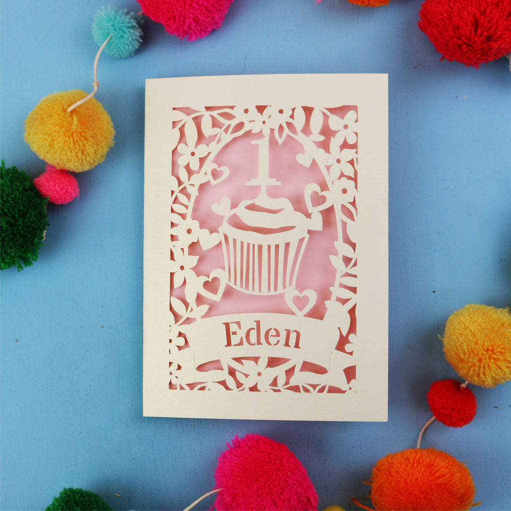Decorative laser cut 1st birthday card with 'Eden' on a blue background with colourful pom-poms
