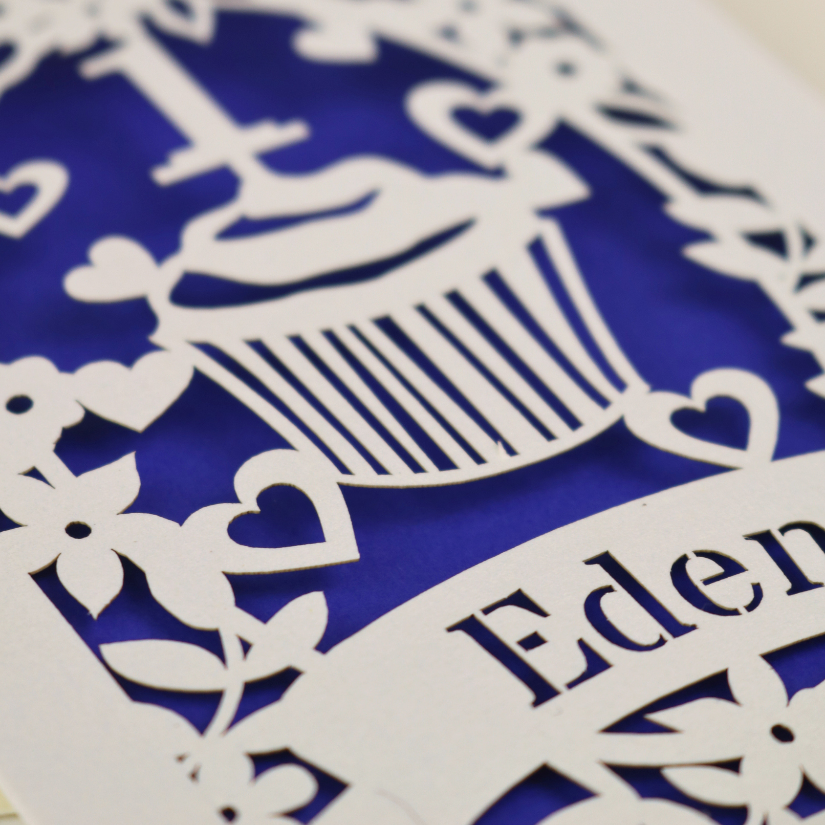 A cream laser cut 1st birthday card with floral border and cupcake design, the number 1 and a banner personalised with a name in cut out stencil font. 