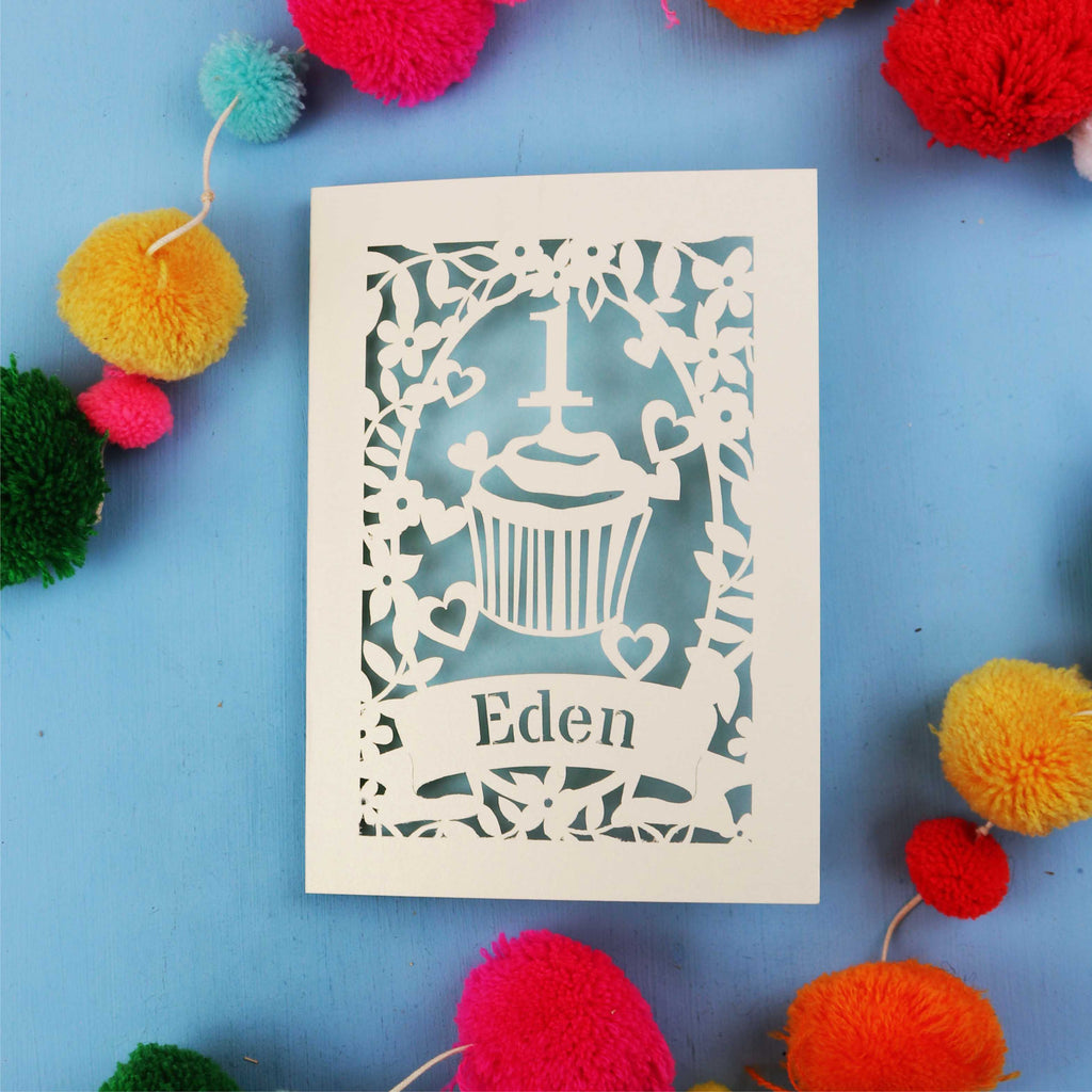 Decorative 1st birthday card with 'Eden' on a blue background with colourful pom-poms