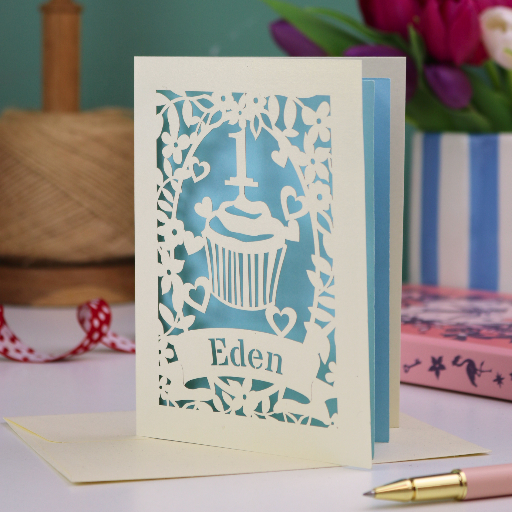 A cream laser cut first birthday card with floral border and cupcake design, the number 1 and a banner personalised with a name in cut out stencil font. 