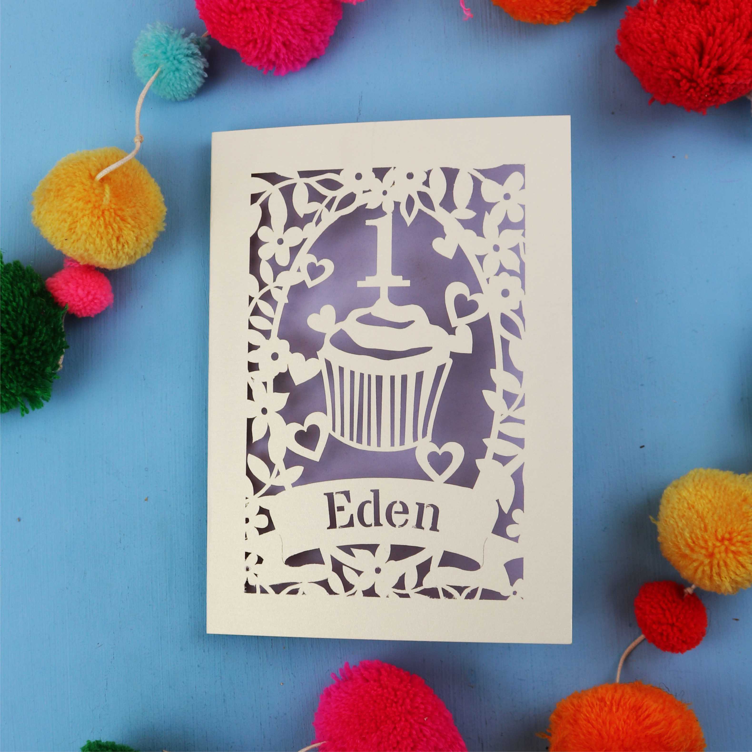 Decorative first birthday card with 'Eden' on a blue background with colourful pom-poms