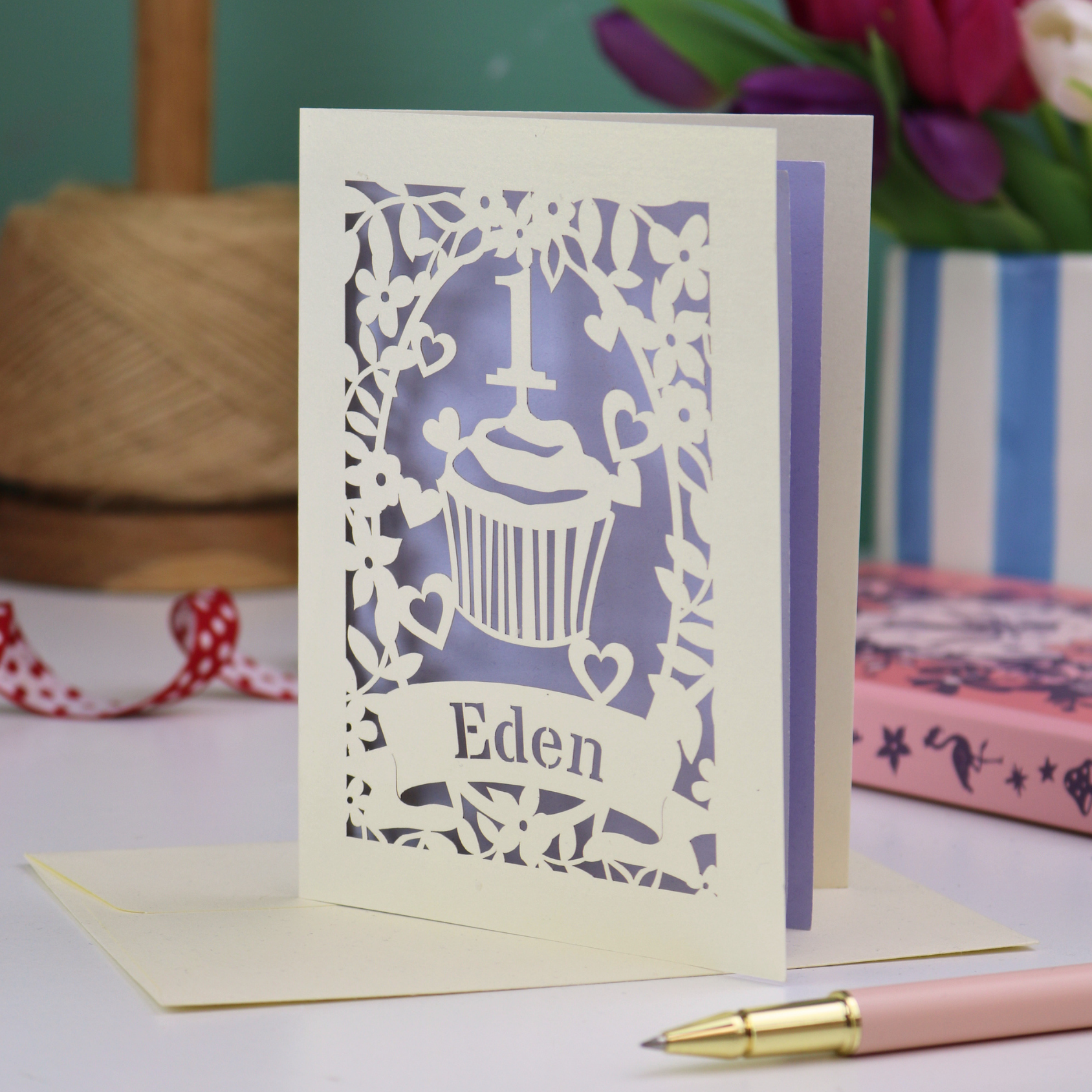 A cream laser cut 1st birthday card with floral border and cupcake design, the number 1 and a banner personalised with a name in cut out stencil font. 