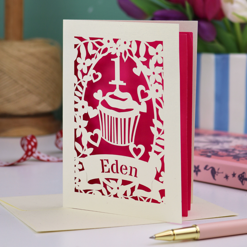 A cream laser cut 1st birthday card with floral border and cupcake design, the number 1 and a banner personalised with a name in cut out stencil font. 