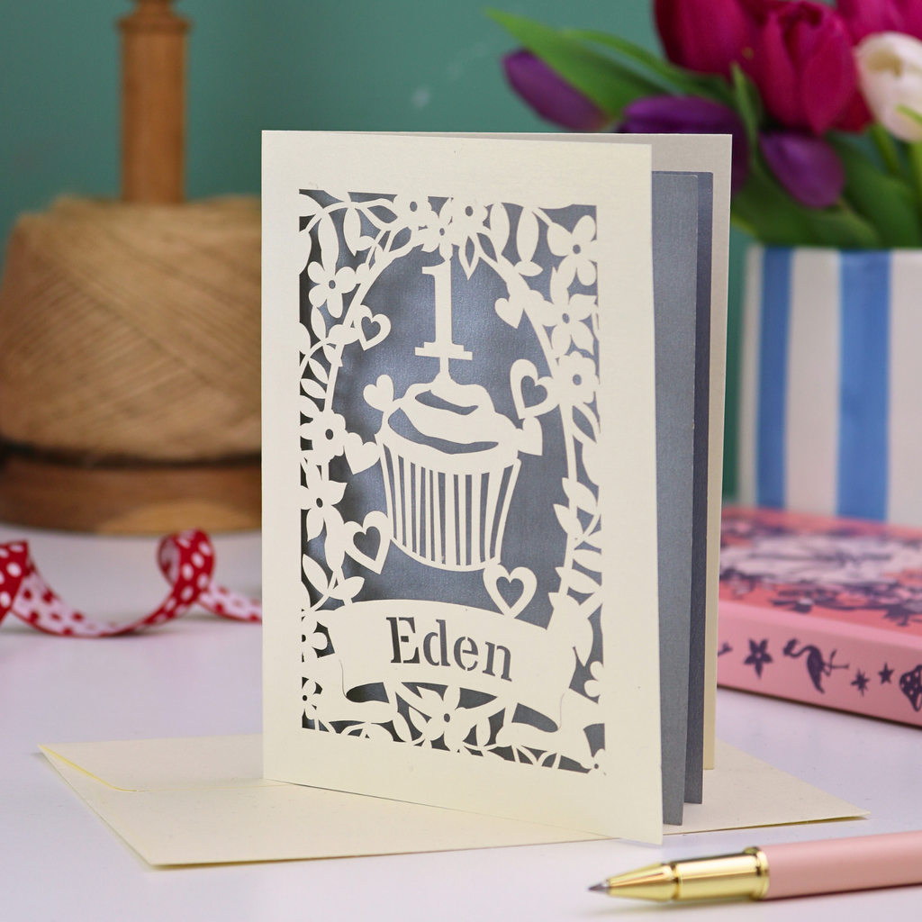 A personalised 1st birthday card with floral border and cupcake design, the number 1 and a banner personalised with a name in cut out stencil font. 