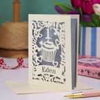 A personalised 1st birthday card with floral border and cupcake design, the number 1 and a banner personalised with a name in cut out stencil font. 