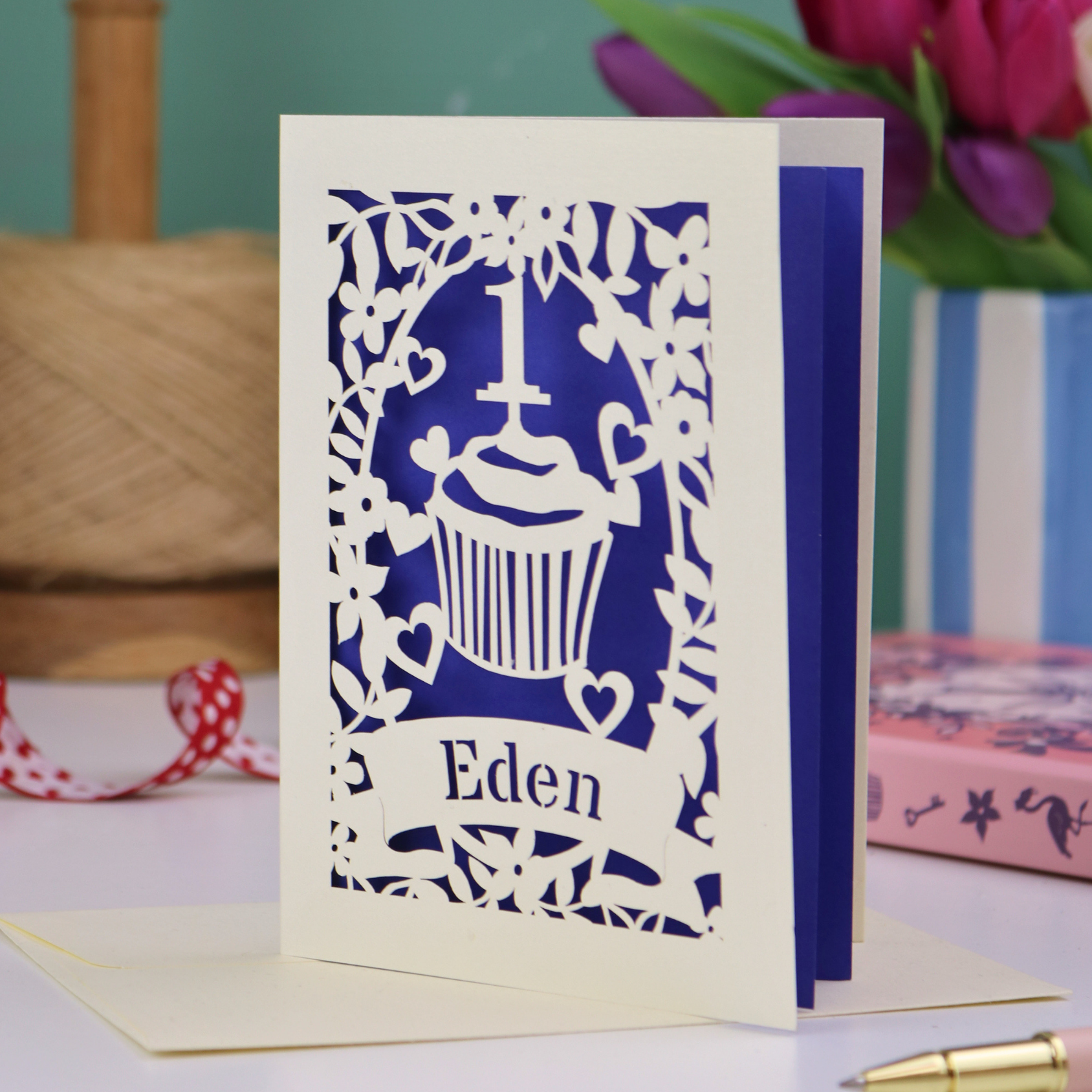 Laser-cut first birthday card with 'Eden' text and decorative design on a table with flowers and thread in the background.