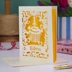 A cream laser cut 1st birthday card with floral border and cupcake design, the number 1 and a banner personalised with a name in cut out stencil font. 