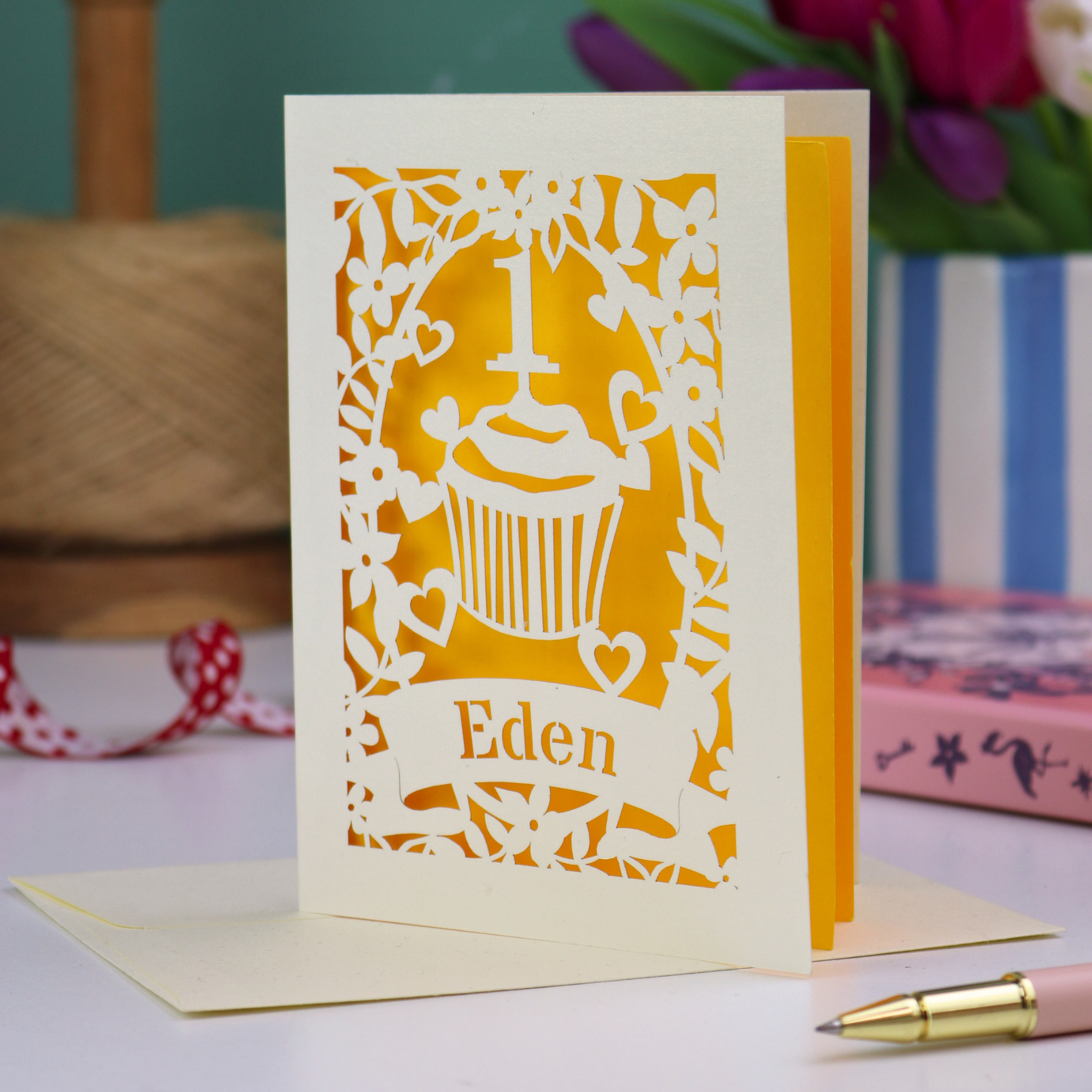 A cream laser cut 1st birthday card with floral border and cupcake design, the number 1 and a banner personalised with a name in cut out stencil font. 