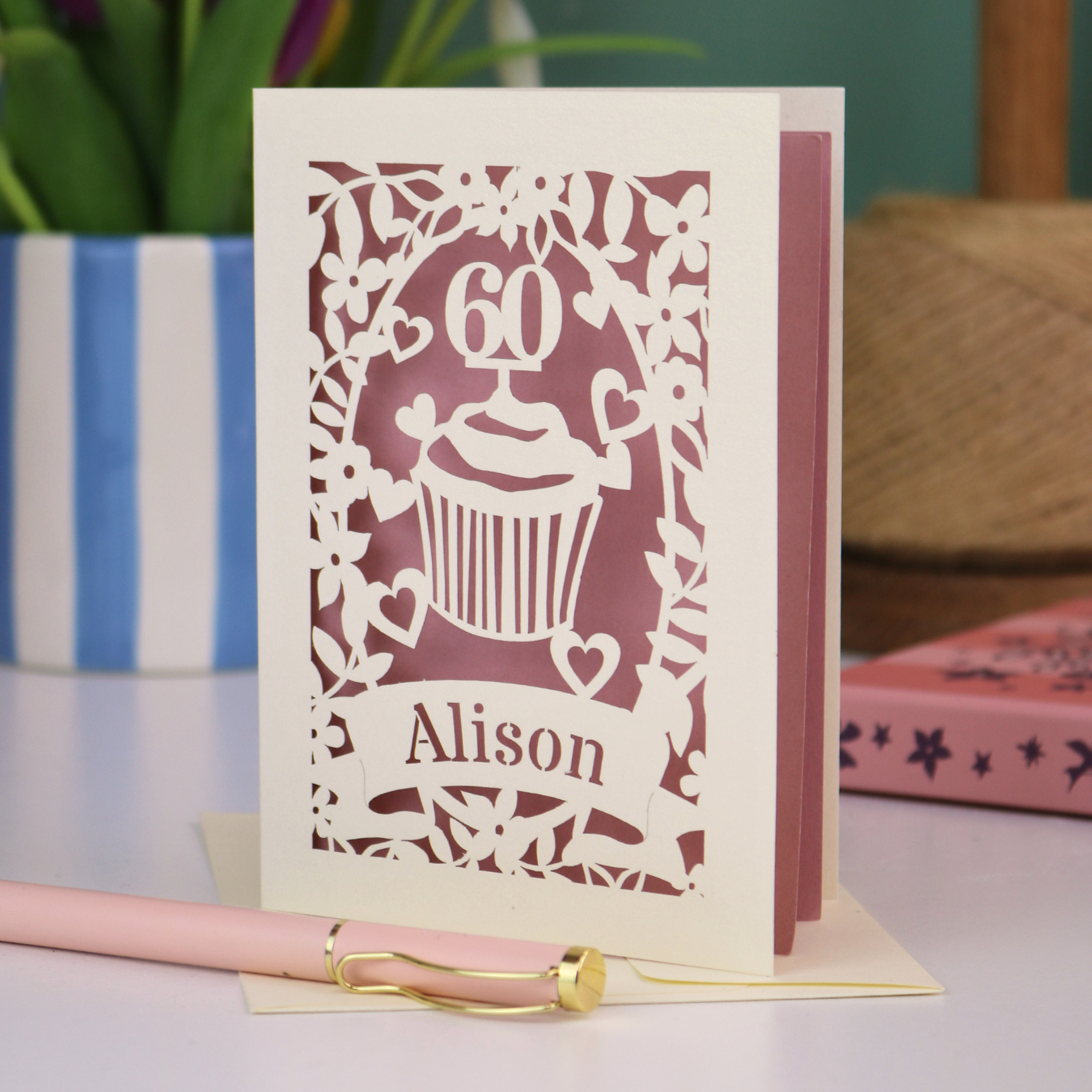 A personalised 60th birthday card with laser cut cupcake design and the age '60', with the name 'Alison' cut out of a banner in a laser cut stencil font. Card is cream and dusky pink