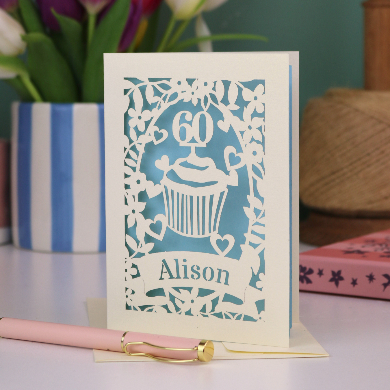 A personalised 60th birthday card with laser cut cupcake design and the age '60', with the name 'Alison' cut out of a banner in a laser cut stencil font. Card is cream and light blue