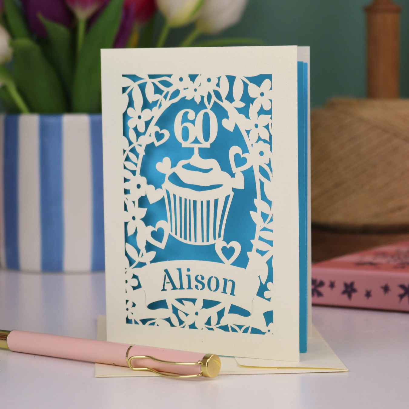 A personalised 60th birthday card with laser cut cupcake design and the age '60', with the name 'Alison' cut out of a banner in a laser cut stencil font. Card is cream and d peacock blue