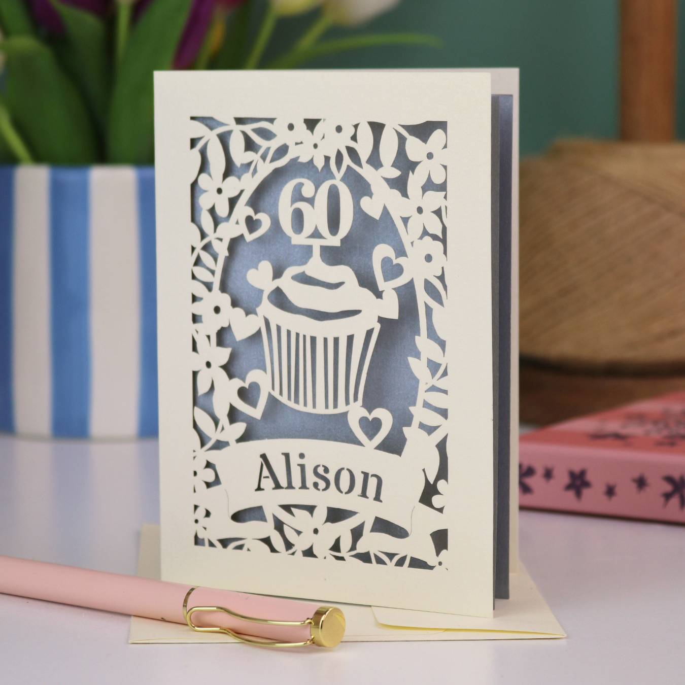 A personalised 60th birthday card with laser cut cupcake design and the age '60', with the name 'Alison' cut out of a banner in a laser cut stencil font. Card is cream and silver
