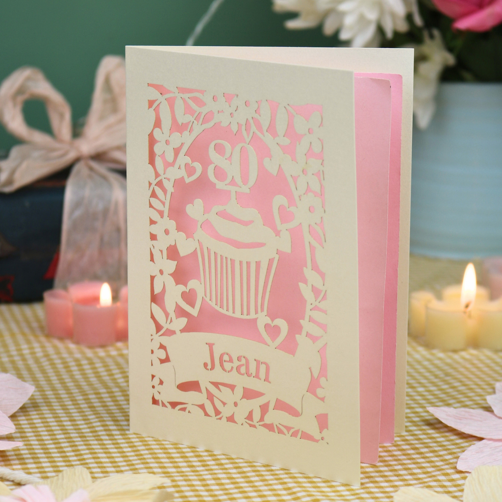 A personalised 80th birthday laser-cut greeting card for Jean, featuring a cream cupcake design with a pink background, displayed on a checked tablecloth with lit candles in the background.