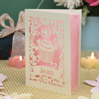 A personalised 80th birthday laser-cut greeting card for Jean, featuring a cream cupcake design with a pink background, displayed on a checked tablecloth with lit candles in the background.