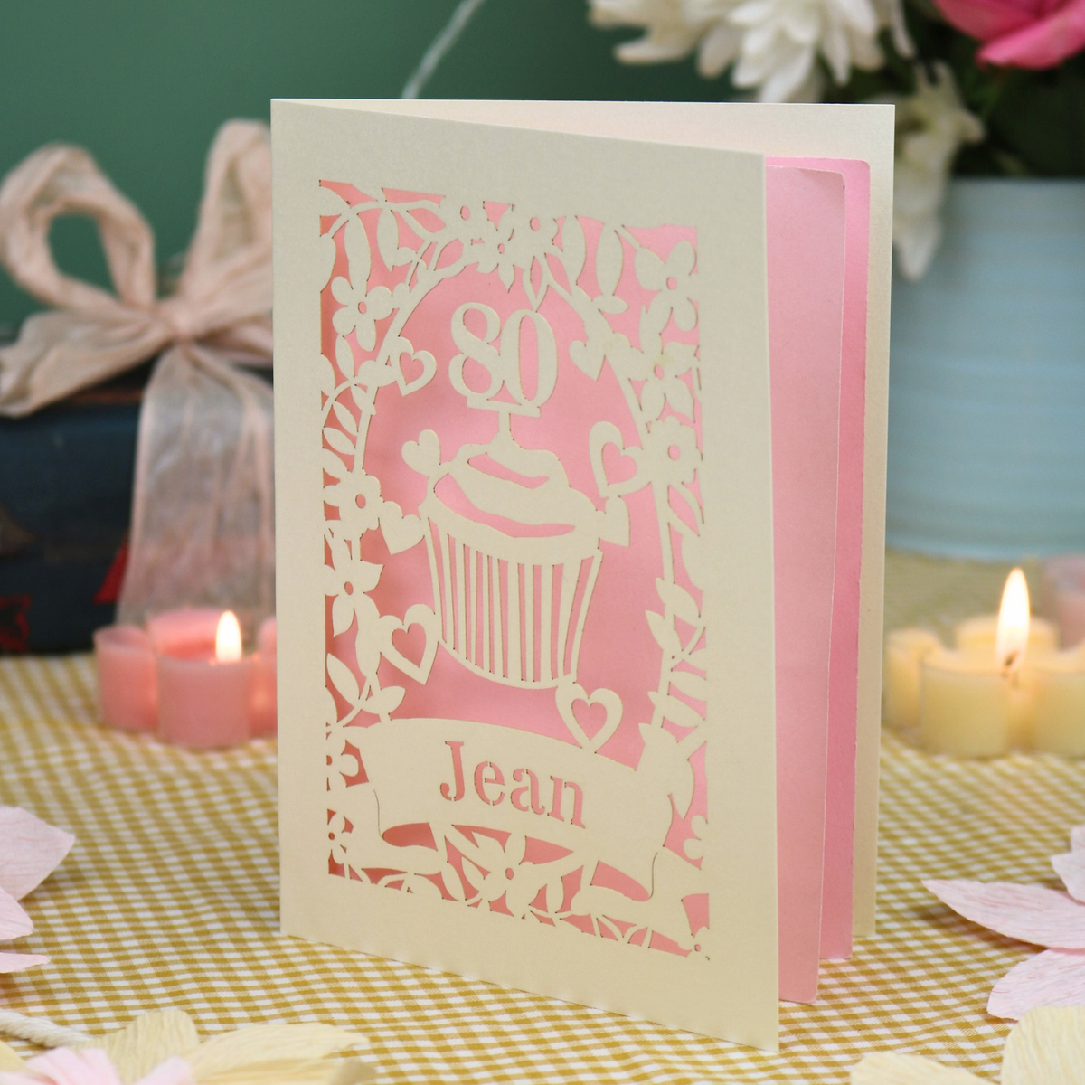 A personalised 80th birthday laser-cut greeting card for Jean, featuring a cream cupcake design with a pink background, displayed on a checked tablecloth with lit candles in the background.