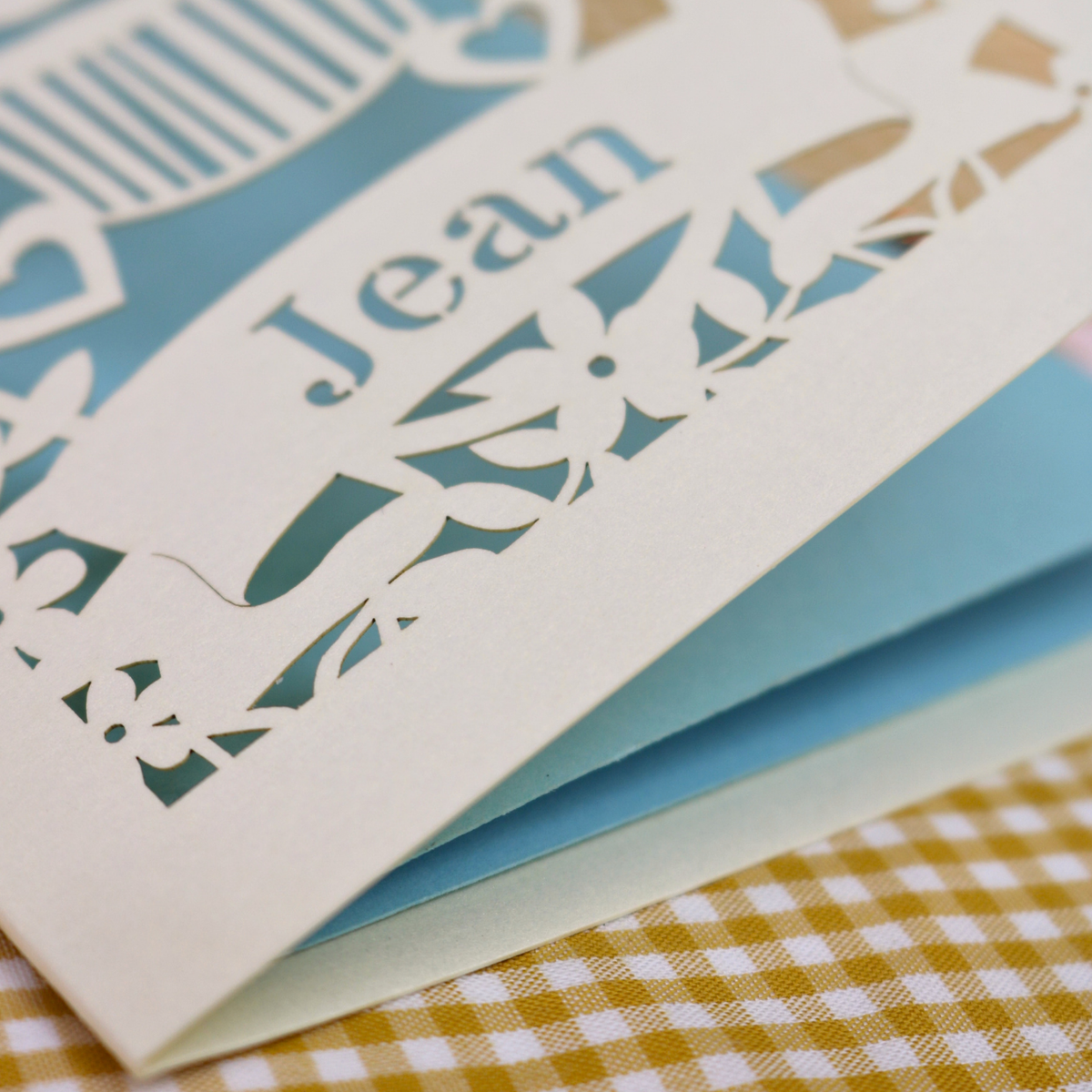 A close-up of a cream-coloured laser-cut  card with the name 'Jean' cut out, layered over a light blue backing, resting on a yellow and white gingham fabric surface.